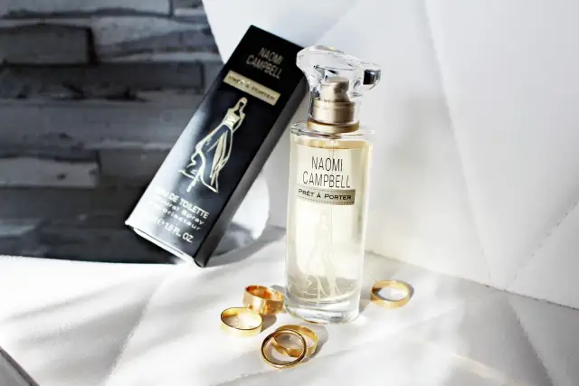 Flakon perfum Naomi Campbell i czarne pudełko z napisem "Eau de Toilette", otoczone złotymi pierścionkami.