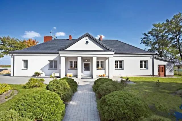 Biały dom z szarym dachem, wejściem z kolumnami i zadbanym ogrodem.