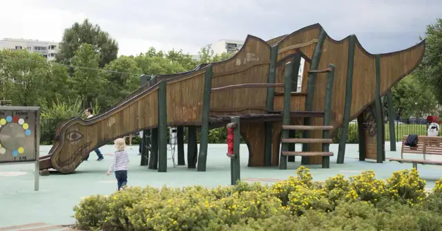 Drewniany plac zabaw w kształcie smoka z dzieckiem i krzaczkami żółtych kwiatów.