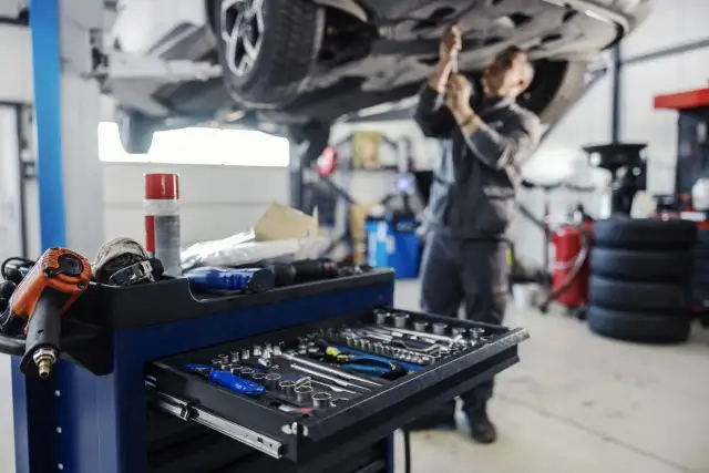 Warsztat samochodowy: mechanik pracuje pod podniesionym autem, wózek z narzędziami na pierwszym planie.