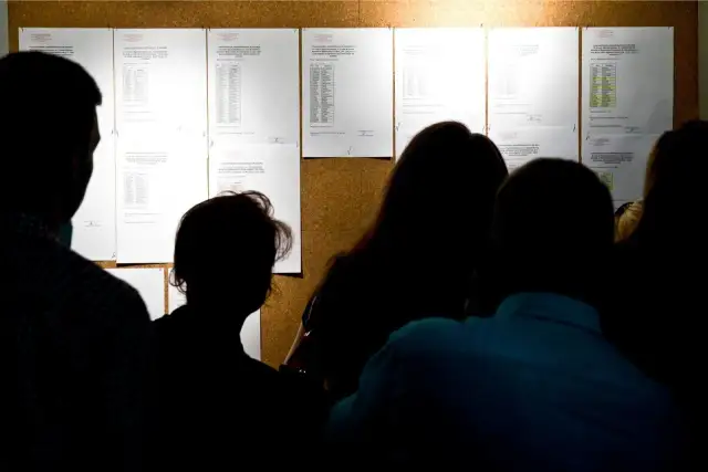 Sylwetki ludzi patrzą na tablicę ogłoszeń z listami wyników rekrutacji do szkół.