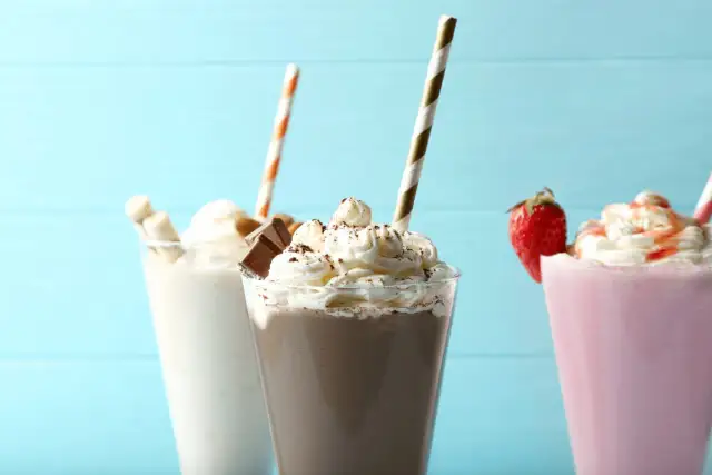 Trzy kolorowe koktajle z bitą śmietaną, słomkami i dekoracjami na niebieskim tle.