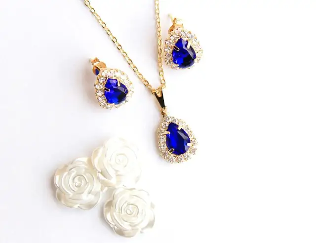 Złoty naszyjnik i kolczyki z niebieskimi kamieniami i cyrkoniami, obok białych róż.