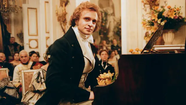 Piotr Adamczyk w historycznej scenie przy fortepianie, otoczony gośćmi w strojach z epoki. Jego role w filmach, serialach i programach zapadają w pamięć.