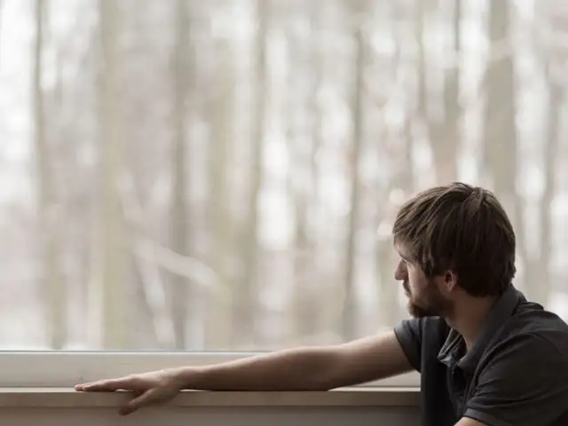 Mężczyzna patrzy przez okno na zimowy las, z ręką opartą na parapecie.