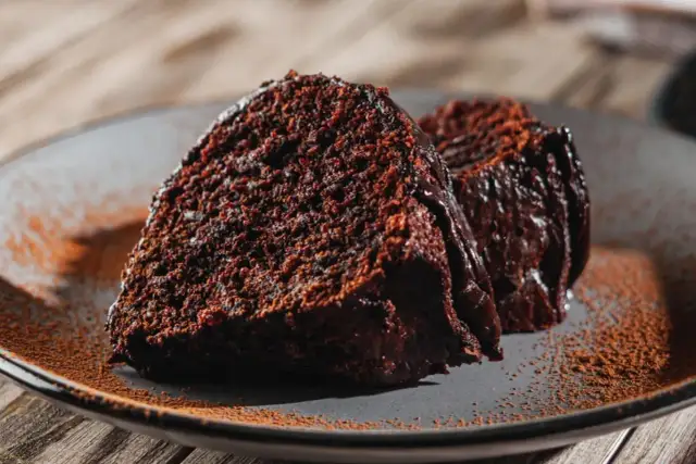 Dwa kawałki wilgotnej babki czekoladowej, posypane kakao, na szarym talerzu.