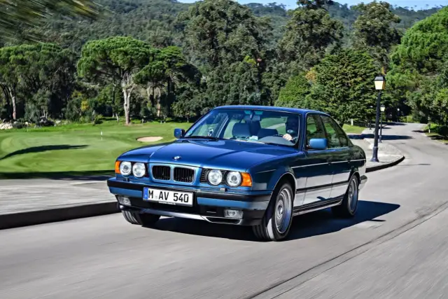 Niebieskie BMW E34 z lat 90. sunie po krętej drodze, mijając malownicze pole golfowe.