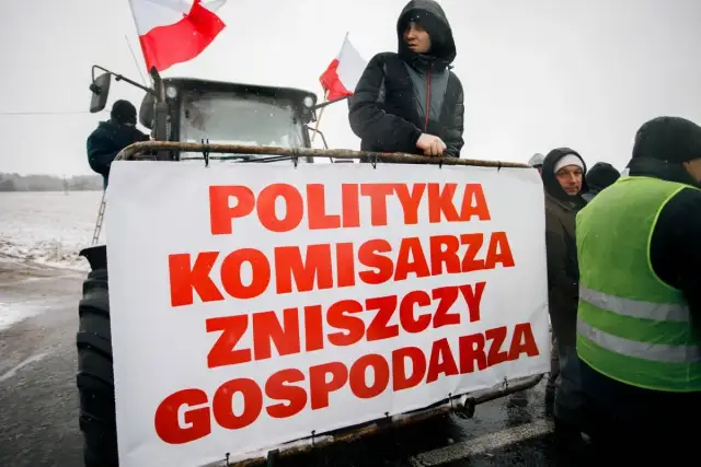 Rolnicy protestują z transparentem "Polityka komisarza zniszczy gospodarza" na tle traktora.