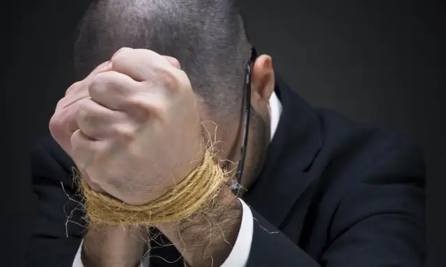 Mężczyzna w garniturze z rękami związanymi sznurkiem, pochylony.