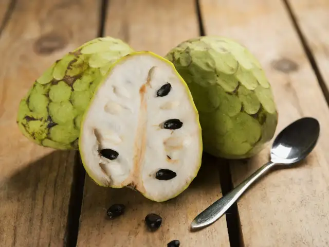 Dwa całe i przekrojone na pół cherimoya owoc z czarnymi pestkami, obok łyżeczka na drewnianym tle.