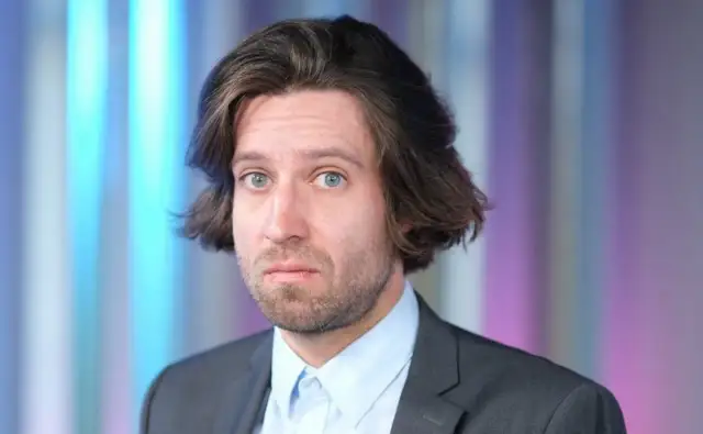 Michał Kempa stand up, w garniturze i jasnej koszuli, z burzą brązowych włosów i niebieskimi oczami, na tle kolorowych pasów.