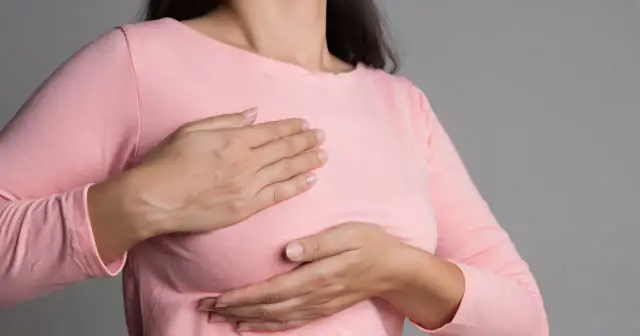 Kobieta w różowej bluzce bada swoje piersi dłońmi.