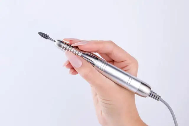 Dłoń z paznokciami trzyma srebrną frezarkę. Zastanawiasz się, jaka moc frezarki do paznokci jest potrzebna do idealnego manicure?