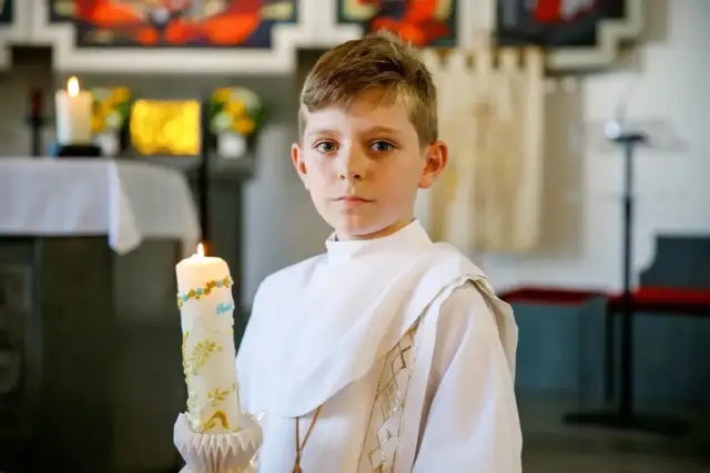 Chłopiec w białej komży trzyma ozdobną świecę podczas komunii świętej.