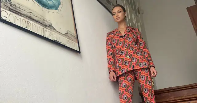 Kobieta w kolorowym, wzorzystym garniturze, przypominającym styl, jak się ubierano w latach 70. Obok plakat teatralny.