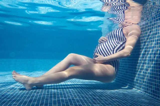 Kobieta w ciąży w pasiastym stroju kąpielowym relaksuje się w basenie. Pływanie w ciąży jakim stylem? Woda koi i odpręża.