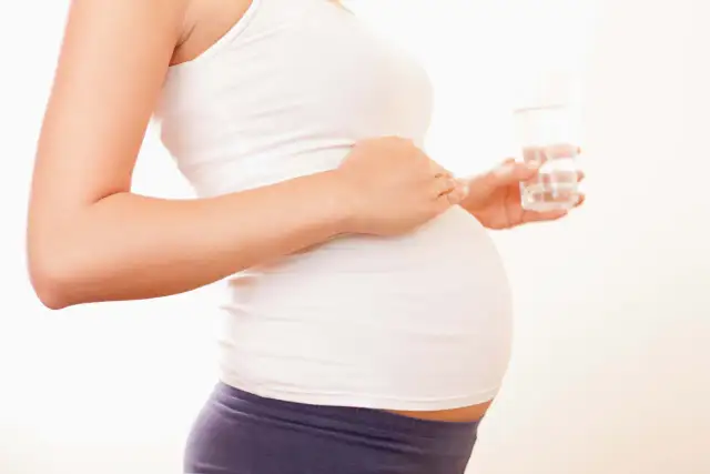 Ciężarna kobieta w białej koszulce trzyma szklankę wody.