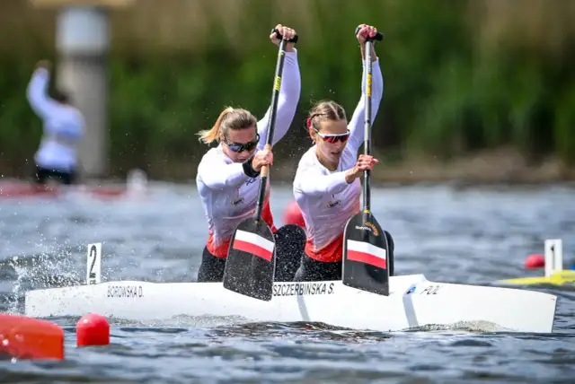 Polki walczą o medal na mistrzostwach świata w kajakarstwie. Dwie zawodniczki w białych strojach z polskimi barwami na wiosłach z determinacją wiosłują w wyścigu.