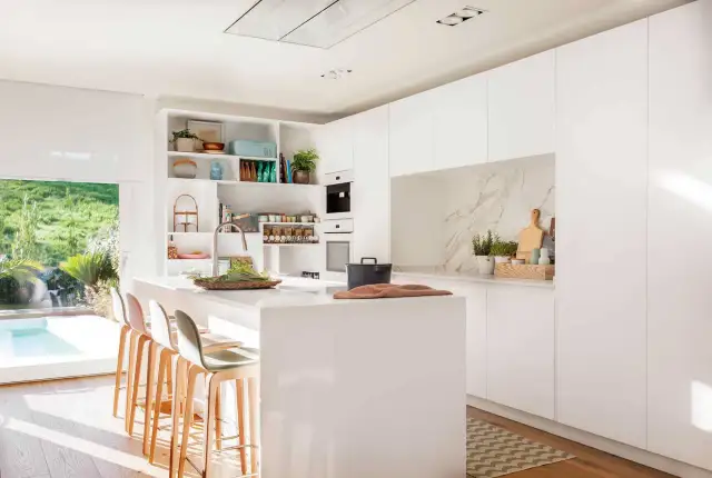 Elegante mueble cocina isla blanco con taburetes, encimera de cuarzo y vistas a un jardín con piscina.