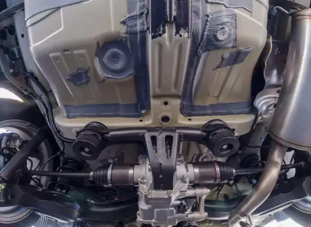 Sprawdź przegub zewnętrzny pod kątem uszkodzeń osłony. Widok podwozia samochodu z widocznym wałem napędowym i elementami zawieszenia.