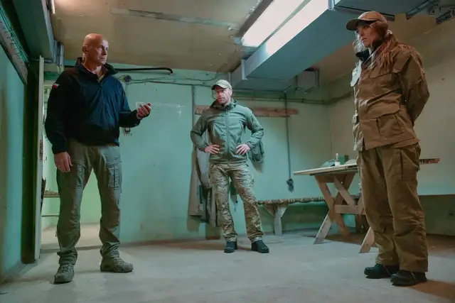 Nasi w mundurach dyskutują w pomieszczeniu. Mężczyzna w czarnej kurtce i dwóch w zielonych strojach taktycznych.