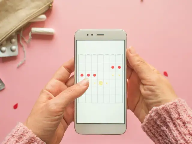 Aplikacja na telefonie pokazuje kalendarz cyklu. Czy można iść na wizytę do ginekologa podczas okresu? Na zdjęciu widać też tampony i tabletki.