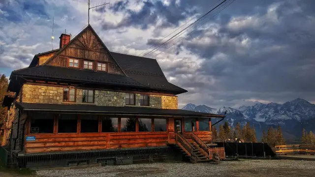 Kamienno-drewniany dom na wierchu, z widokiem na ośnieżone szczyty gór.