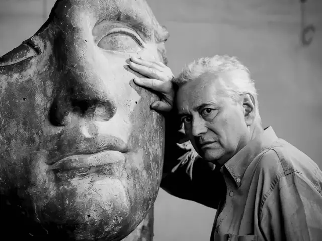 Czarno-białe zdjęcie artysty Igora Mitoraja opierającego się o fragment swojej rzeźby przedstawiającej twarz.