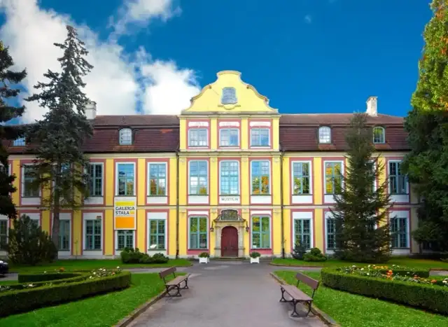 Żółty budynek Muzeum Narodowego w Gdańsku z czerwonym dachem i zielonymi oknami, otoczony ogrodem.