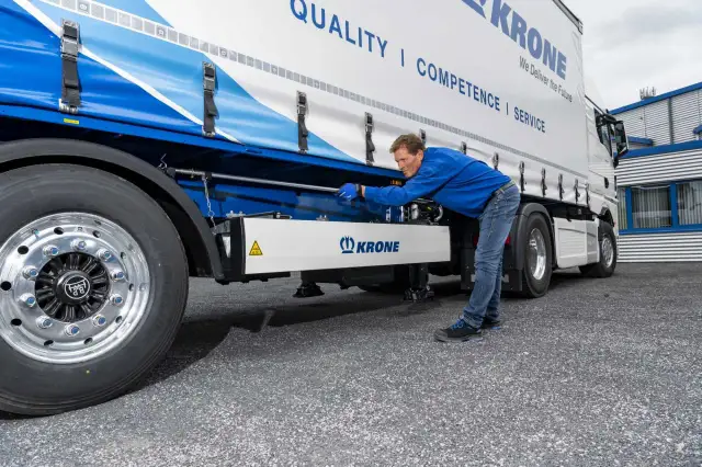 Mężczyzna w niebieskiej kurtce pochyla się nad naczepą Krone, szukając bezpieczników.