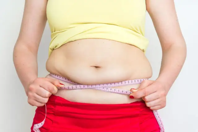 Kobieta mierzy obwód talii centymetrem. Widoczny jest odstający brzuch poniżej pępka, co może świadczyć o nadwadze.