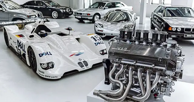 Silnik V12 BMW Motorsport obok białego bolidu wyścigowego i kilku klasycznych BMW.