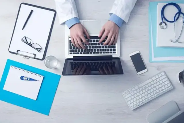 Lekarz wpisuje dane na laptopie, pokazując, jak zamówić leki na receptę przez internet. Na biurku leżą też stetoskop, okulary i telefon.