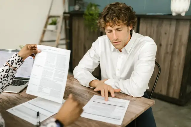 Mężczyzna w białej koszuli analizuje dokumenty z kobietą przy drewnianym stole.