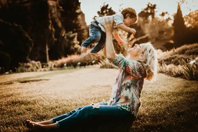 Mama rzuca dzieckiem w górę podczas śmiesznej sesji rodzinnej w parku. Słońce zachodzi, tworząc ciepłą, złotą poświatę.