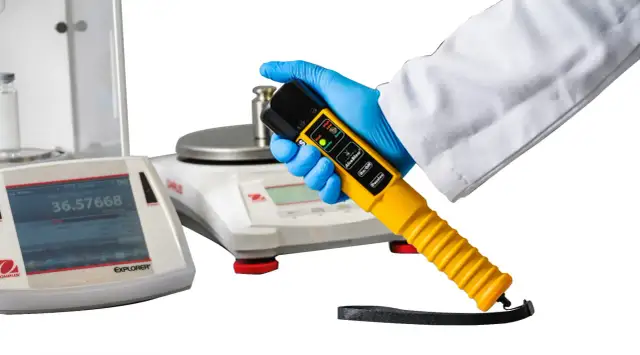 Ręka w niebieskiej rękawiczce trzyma żółty alkomat. W tle waga laboratoryjna.