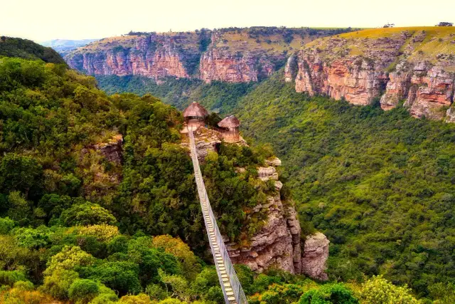 Most zwodzony do chat na skale w górach przylądkowych. Zielone zbocza i skalne klify tworzą malowniczy krajobraz.