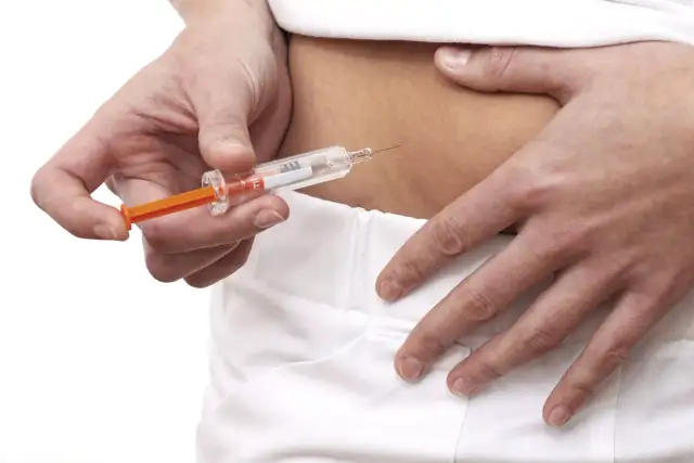 Opiekun medyczny może robić zastrzyki. Dłoń z igłą strzykawki zbliża się do skóry brzucha.