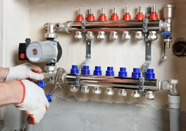 Instalator w rękawiczkach dokręca kluczem pompę obiegową do rozdzielacza ogrzewania podłogowego.