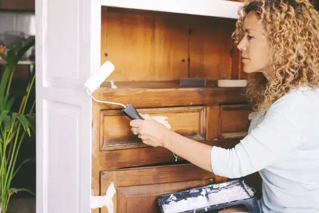 Kobieta maluje wałkiem mebel z okleiny na biało.