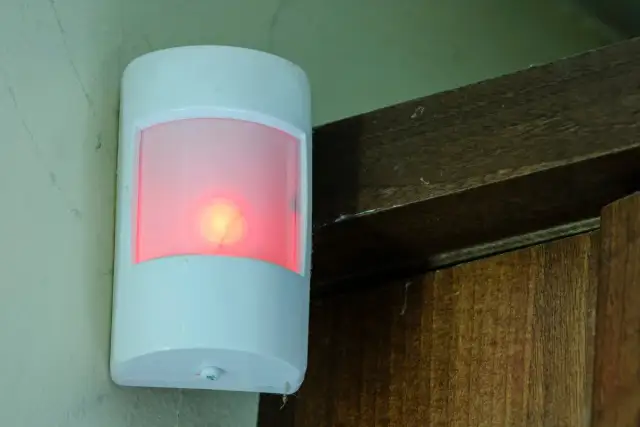 Biały czujnik ruchu z czerwoną diodą, zamontowany na ścianie obok drewnianej framugi. Pokazuje, jak działa czujnik ruchu.