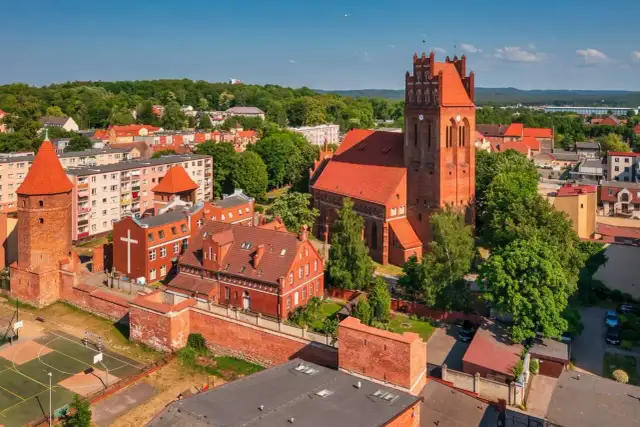 Zamek krzyżacki w Lęborku, z charakterystyczną wieżą i ceglaną architekturą, otoczony zielenią i zabudowaniami miejskimi.