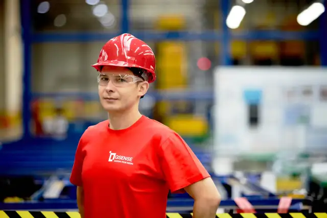 Mężczyzna w czerwonym kasku i okularach ochronnych, w czerwonej koszulce z logo firmy, na tle hali produkcyjnej.