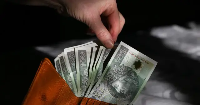 Dłoń wyciąga banknot 100 zł z brązowego portfela. Pieniądze, które mogą być przeznaczone dla kogo czternasta emerytura.
