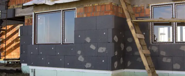 Budowa domu w toku, trwa ocieplanie ścian zewnętrznych styropianem. Widać okna i fragment rusztowania.