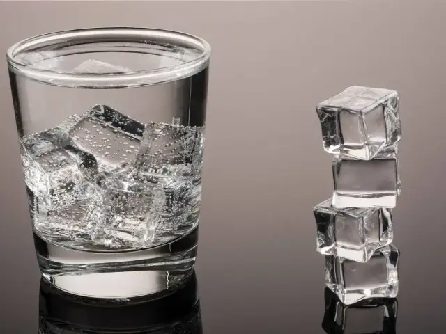 Szklanka wody z lodem i wieża z kostek lodu na szarym tle.