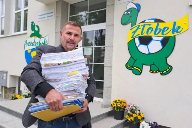 Mężczyzna z dużą stertą dokumentów przed żłobkiem z logo żółwia.