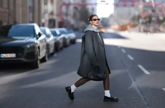 Kobieta w szarym płaszczu i białych skarpetkach z czarnymi mokasynami przechodzi przez ulicę.