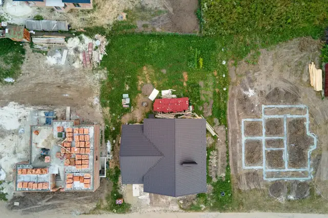 Widok z lotu ptaka na plac budowy z fundamentami, częściowo zbudowanym domem i materiałami budowlanymi.
