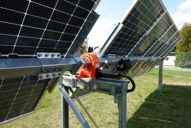 Mechanizm tracker pv z pomarańczową przekładnią i siłownikiem, obracający panele słoneczne na metalowej konstrukcji.